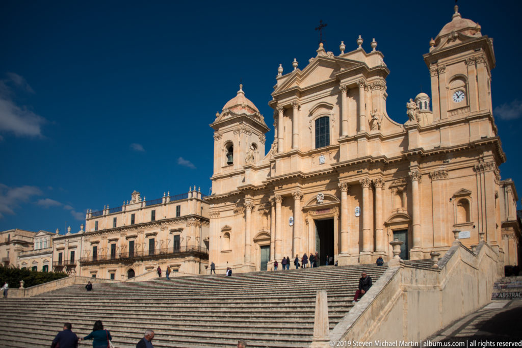 Wide view of Noto Cathedral above its broad staircase, with twin towers, visitors, and an adjoining palace.