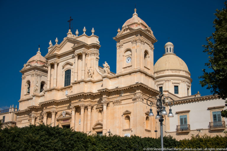 Angled view of Noto Cathedral with twin bell towers, Baroque façade, clock faces, and dome against blue sky.