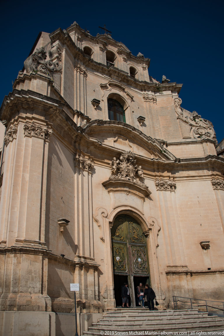 Oblique low-angle view of a monumental Baroque church façade with giant pilasters, sculpted doorway, and visitors at the entrance.