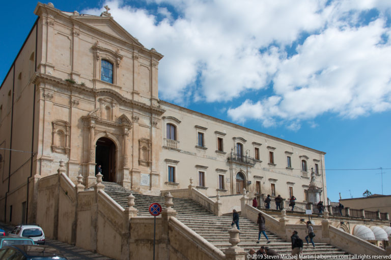 Oblique view of a Baroque church atop broad stairs beside a long historic building with balconies.