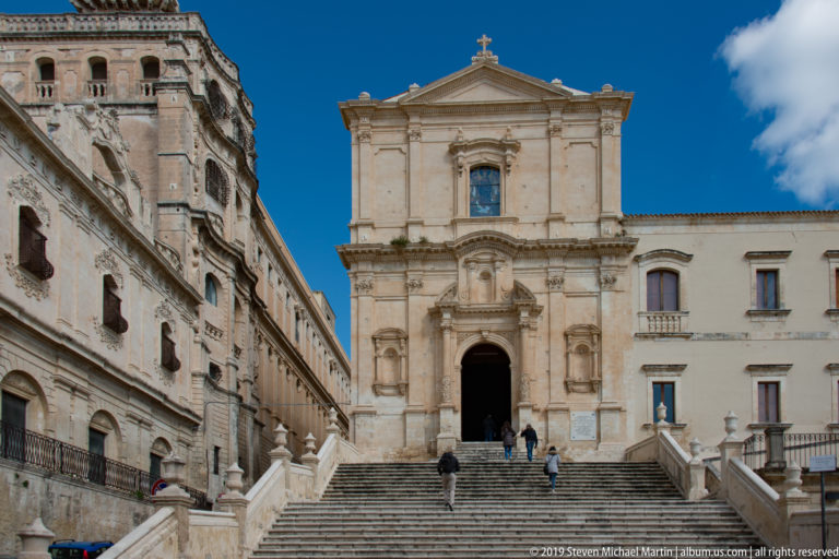 Baroque church with tall central portal and pediment above a broad stone staircase, with visitors climbing the steps.