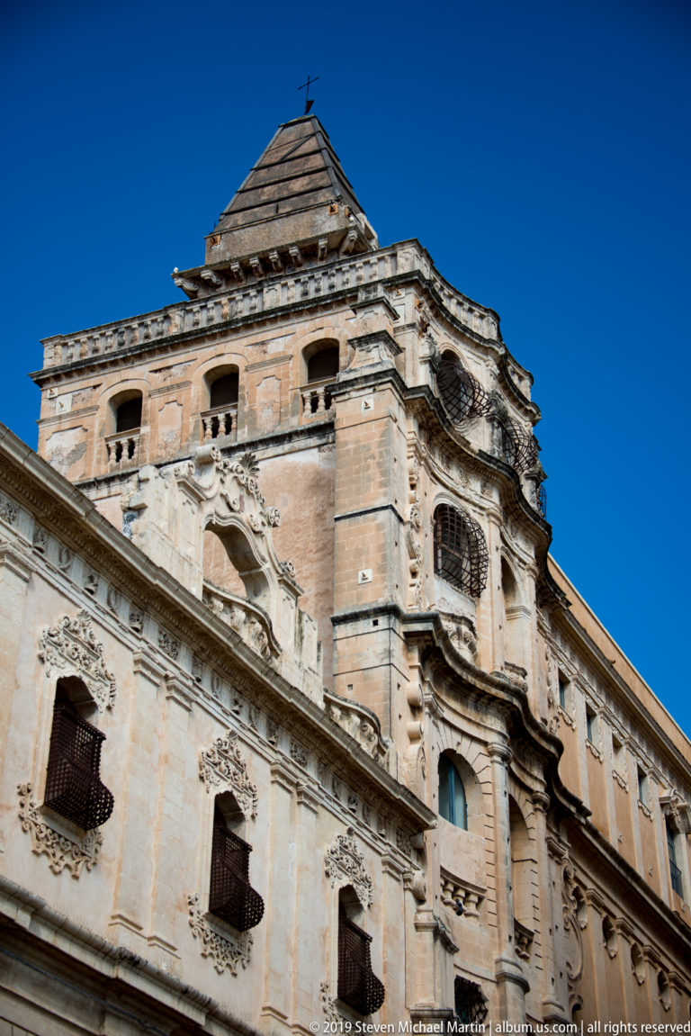 Low-angle view of a weathered Baroque stone building with carved windows, balconies, cornices, and a tall rooftop pavilion.