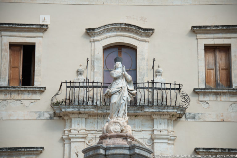Close view of a draped Virgin Mary statue on a pedestal aligned with a wrought-iron balcony behind.