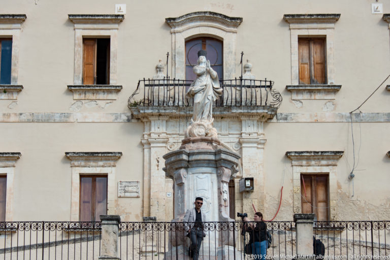 Virgin Mary statue on a pedestal in front of an iron balcony and historic building, with pedestrians below.