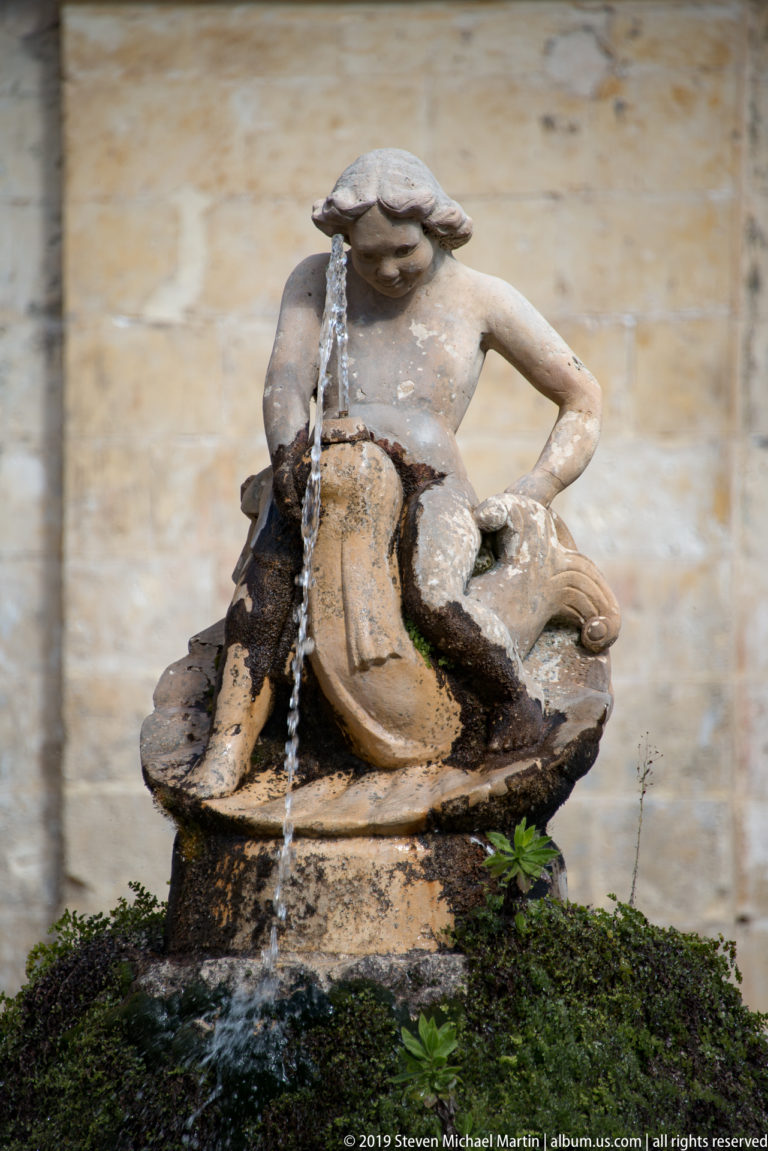 Weathered reclining female stone figure with water streaming over the sculpture and moss growing around its base.
