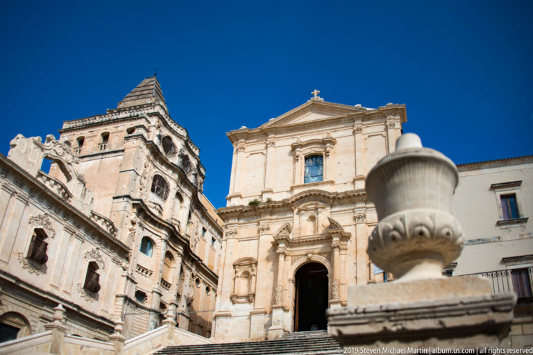 Baroque church façade at the top of broad stone stairs beside a large historic building under deep blue sky.