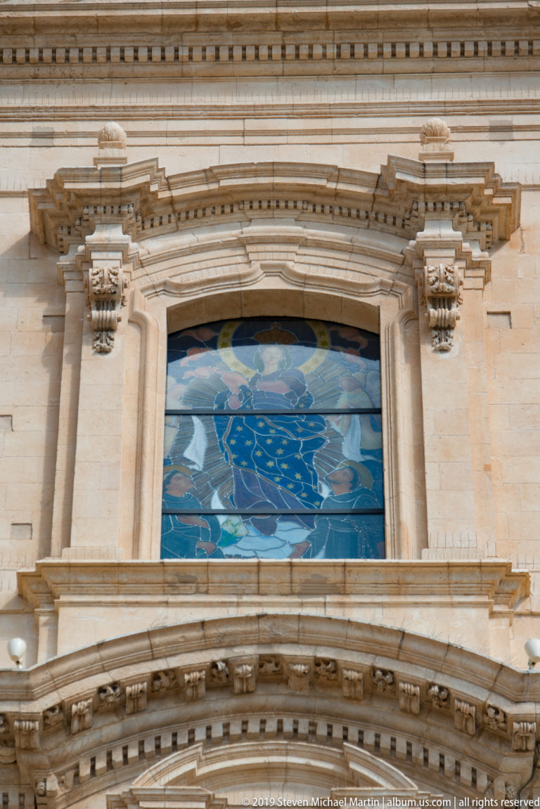 Blue Marian stained-glass window set within an elaborate pale limestone Baroque surround on a church façade.