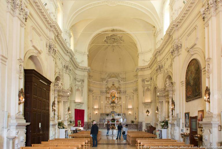 Bright Baroque church nave with wooden pews, side altars, paintings, visitors, and sanctuary at the far end.