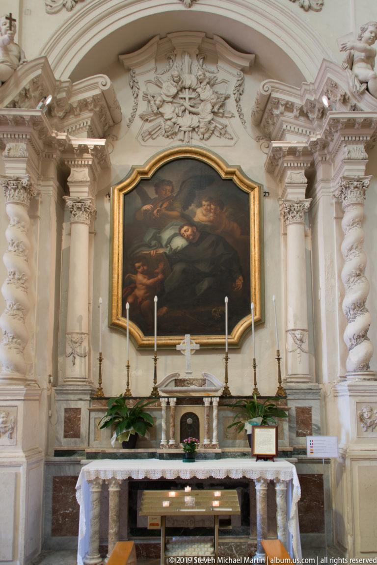 Ornate side altar with twisted columns, white stucco relief, gold trim, candlesticks, and a dark religious painting.