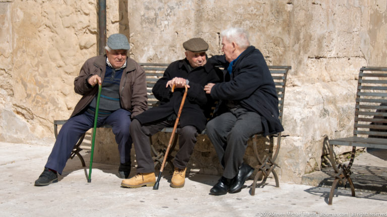 Three elderly men in dark clothing sit talking on metal chairs beside a rough stone wall, two holding walking sticks.