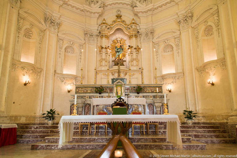 White-and-gold church sanctuary with marble altar, central devotional statue, candlesticks, and symmetrical stucco decoration.