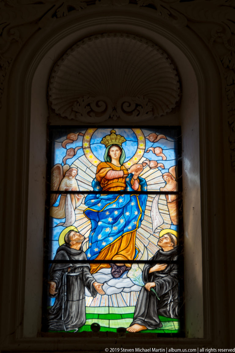 Illuminated stained-glass window showing a crowned Virgin in blue above two kneeling religious figures.