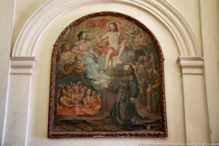 Arched religious painting with Christ above clustered figures and a dark-robed kneeling friar at lower right.