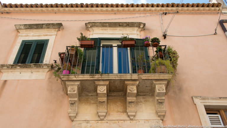 Small stone balcony with carved supports, potted plants, green shutters, and a central glass door on a peach façade.