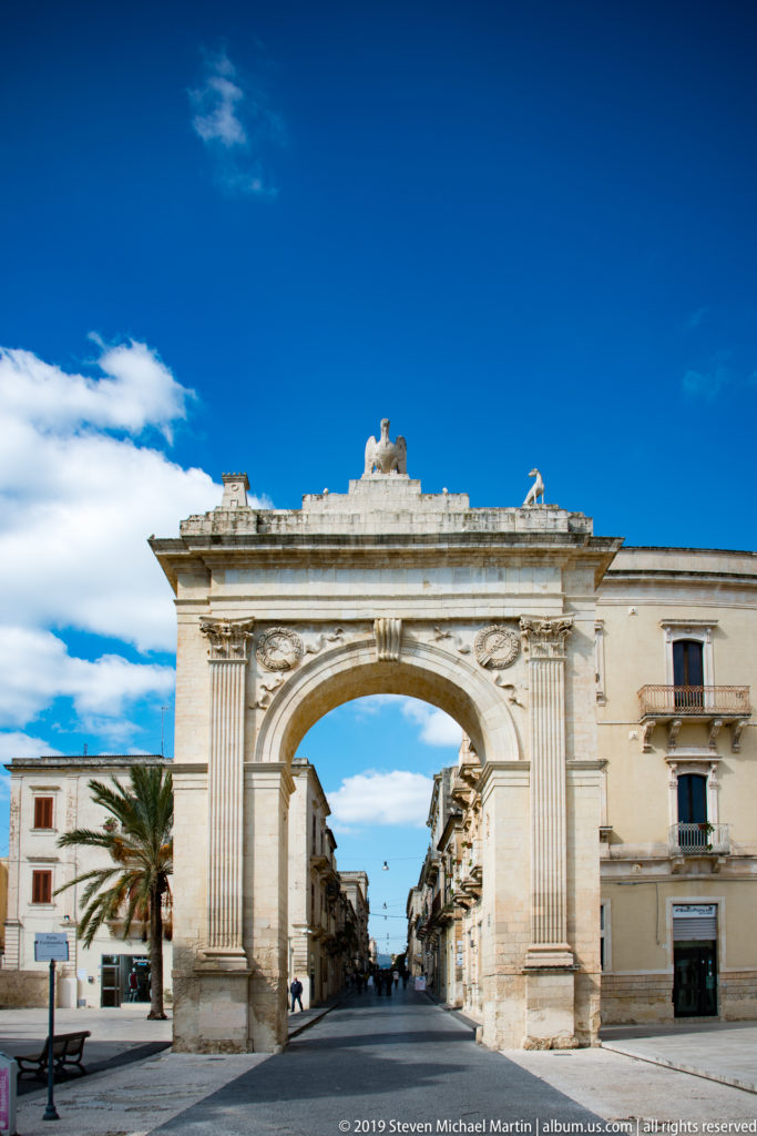 Frontal view of Porta Reale, a pale stone triumphal arch opening onto a straight street under blue sky.