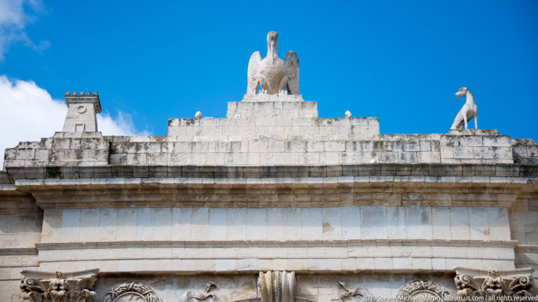 Stone bird sculpture and smaller rooftop figure above the cornice of Porta Reale against a deep blue sky.