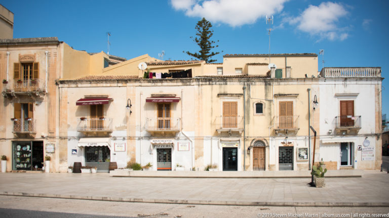 Weathered cream and ochre buildings with balconies, shutters, shops, and roof terraces facing a broad paved square.
