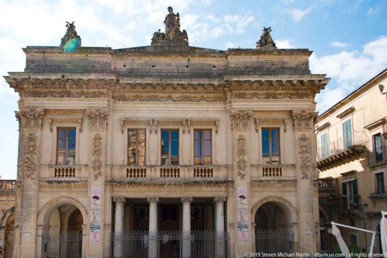 Symmetrical limestone palazzo with three lower arches, ornate upper windows, pilasters, balustrades, and rooftop sculptures.