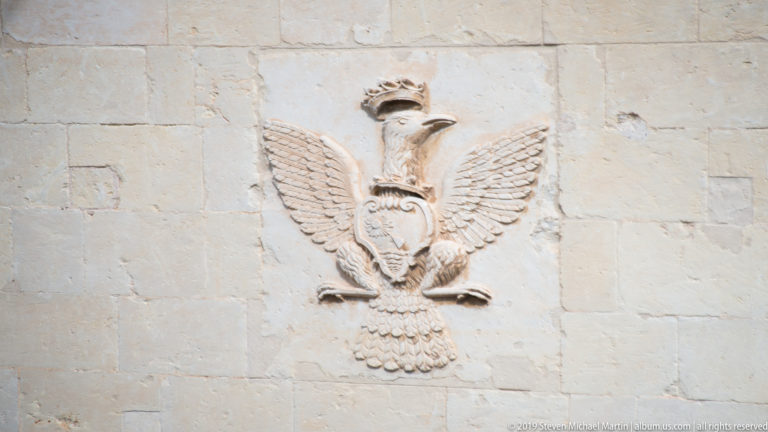 Crowned eagle relief with spread wings and a shield-like emblem carved into a pale limestone wall.