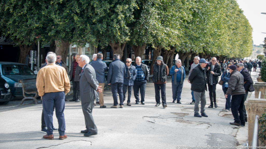 Groups of men stand talking along a tree-shaded pedestrian area with benches and parked vehicles nearby.
