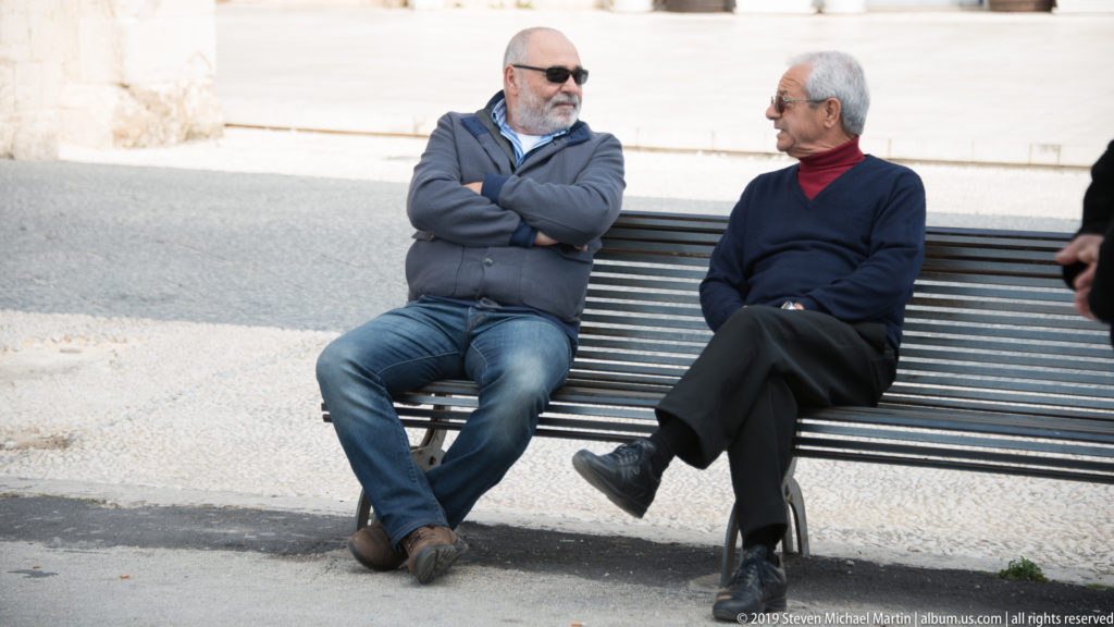 Two older men seated on a slatted bench facing one another in conversation beside pale stone paving.