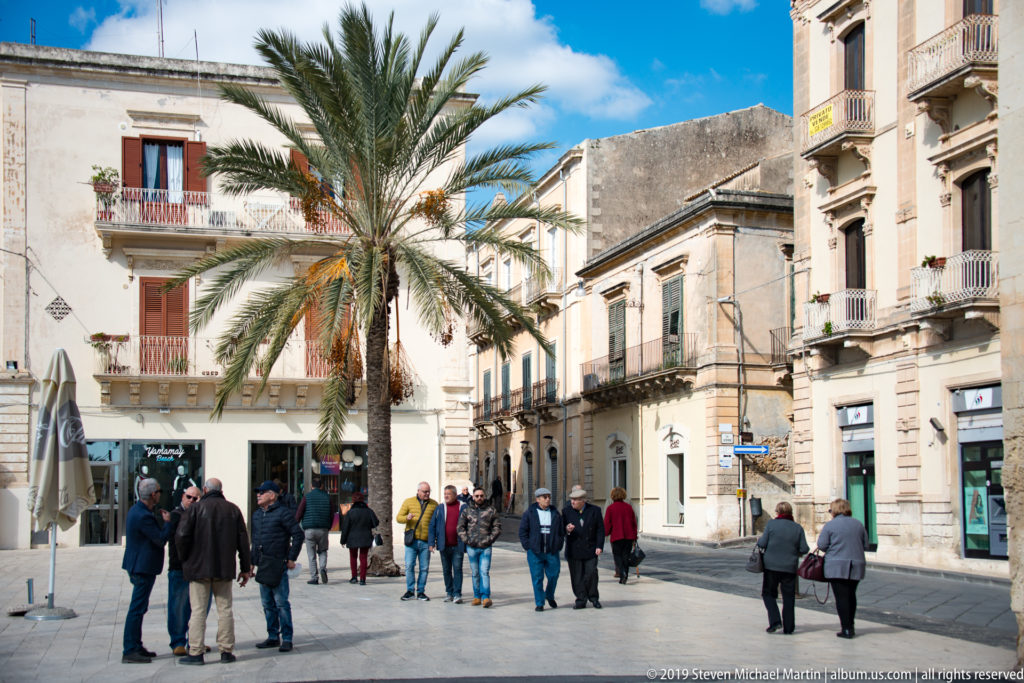 Pedestrians gather beneath palm trees beside pale historic buildings with balconies and shuttered windows in Noto.