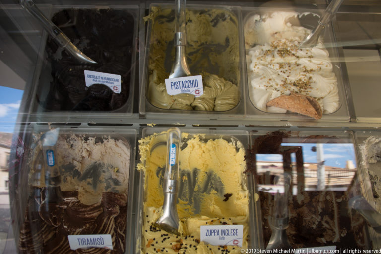Gelato case with several open tubs, serving spatulas, and labels including pistachio and tiramisu.