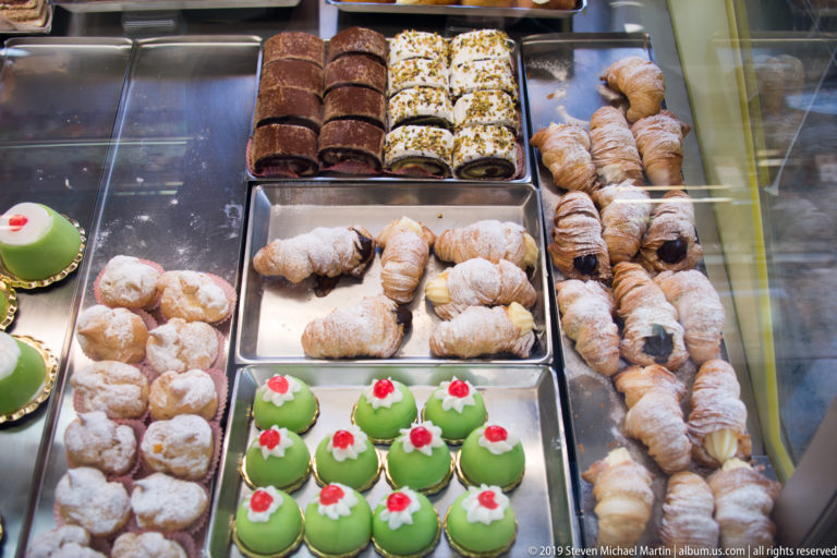 Glass bakery case containing trays of powdered pastries, rolled sweets, green domes, and filled pastries.