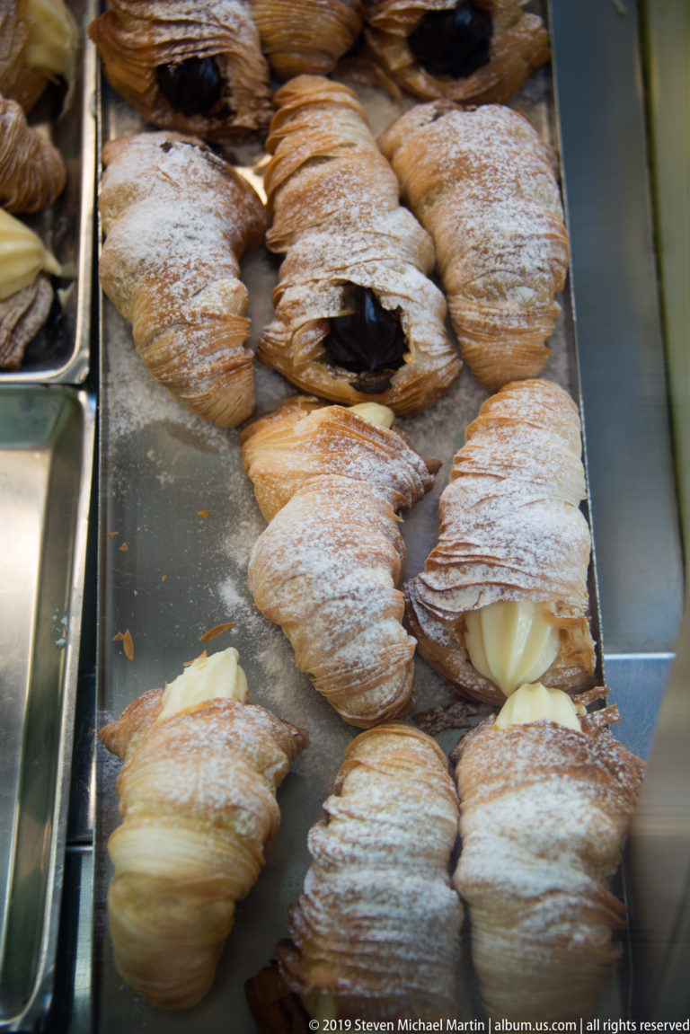 Powdered pastries filled with pale cream and dark chocolate arranged in rows on metal bakery trays.