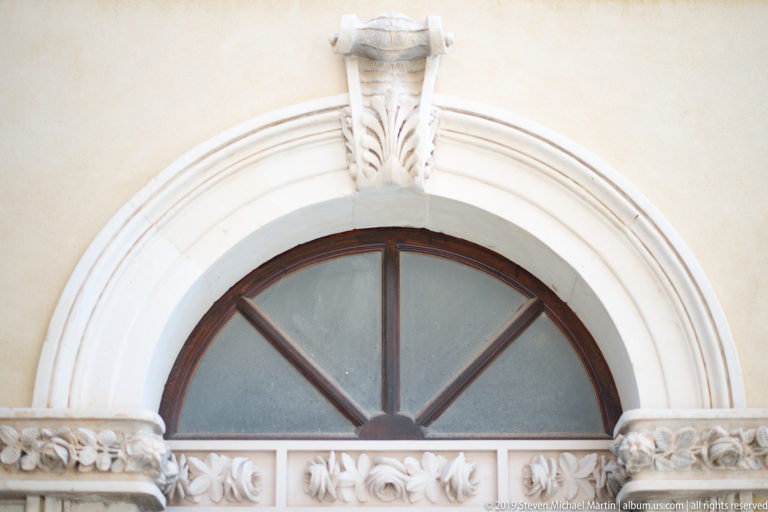Semicircular dark-wood window framed by pale carved stone, with a keystone above and floral relief band below.