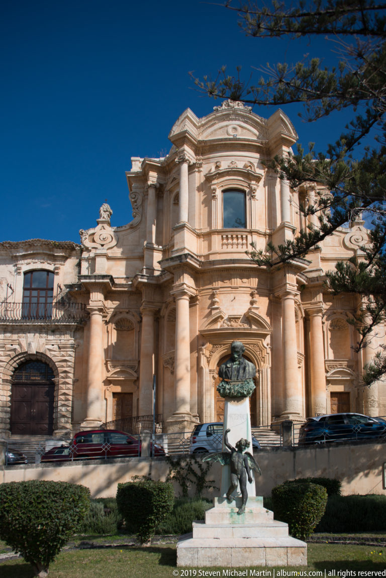 Curving Baroque church façade behind a foreground monument, clipped shrubs, parked cars, and a deep blue sky.