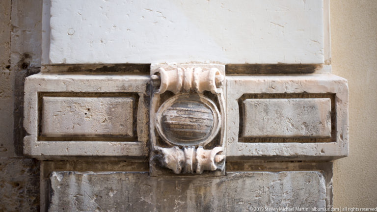 Weathered circular stone medallion with scroll-like sides centered in a rectangular band of pale masonry blocks.