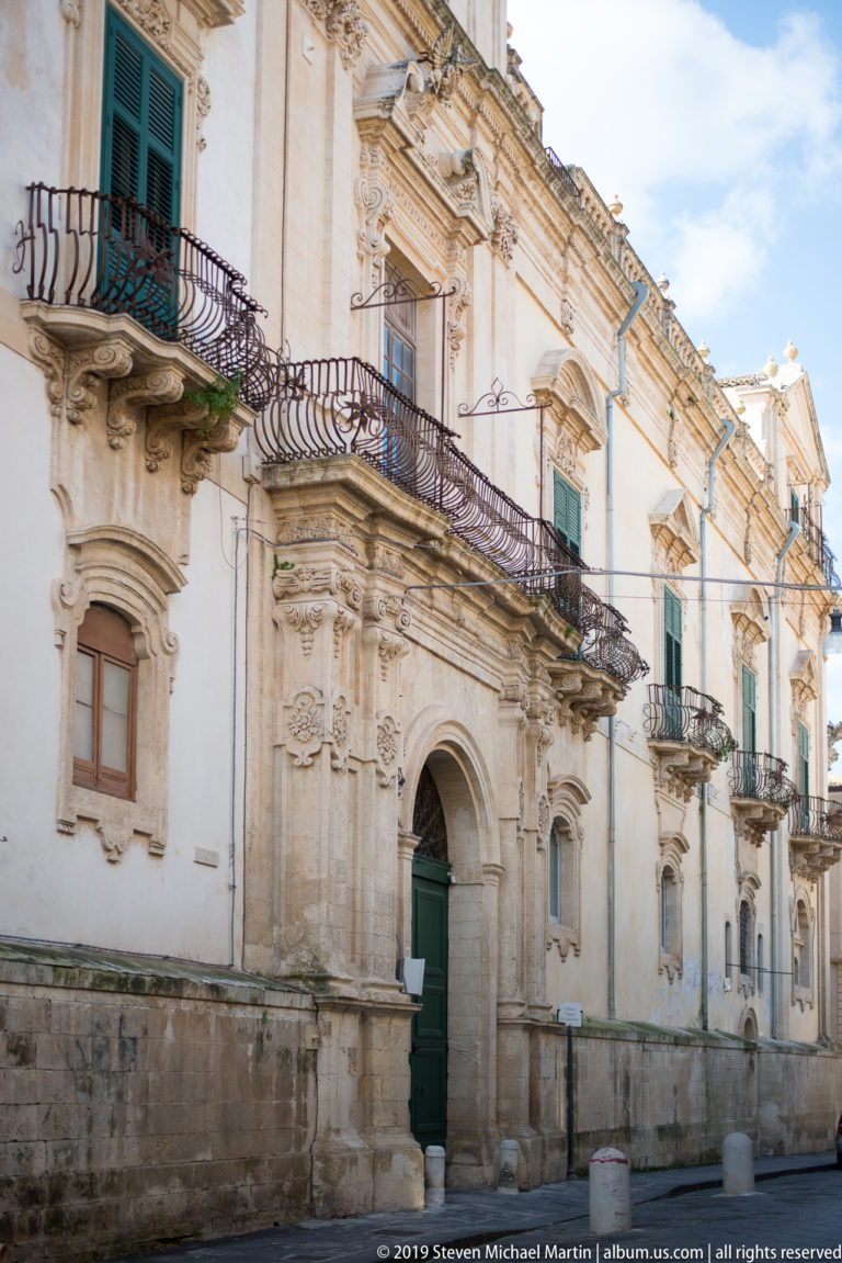 Pale Baroque palace façade with arched doorway, carved window surrounds, projecting iron balconies, and green shutters.