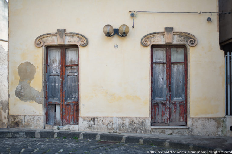 Two weathered red-brown wooden doors in carved pale-stone surrounds on a worn cream plaster wall.