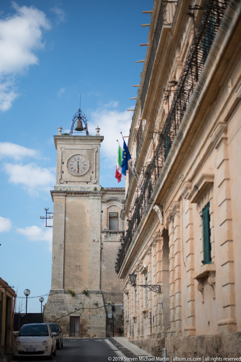 Stone clock tower beside a long historic civic façade with balconies and Italian and European Union flags.
