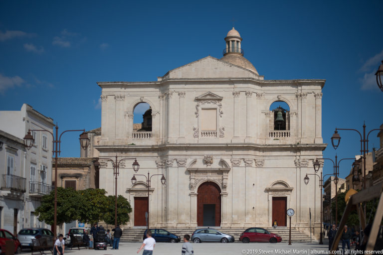 Pale Baroque church façade with three ground-level doors, two open upper arches, small roof lantern, cars, and pedestrians.