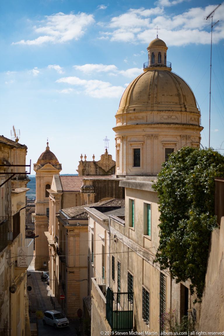 Large tan masonry dome and lantern rising above a narrow street lined with pale historic buildings in Noto.