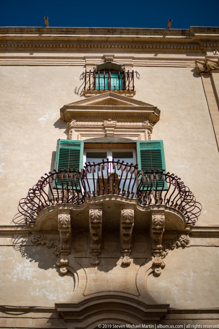 Low-angle view of a stone balcony with curved wrought-iron railing and green shutters on a pale historic façade.