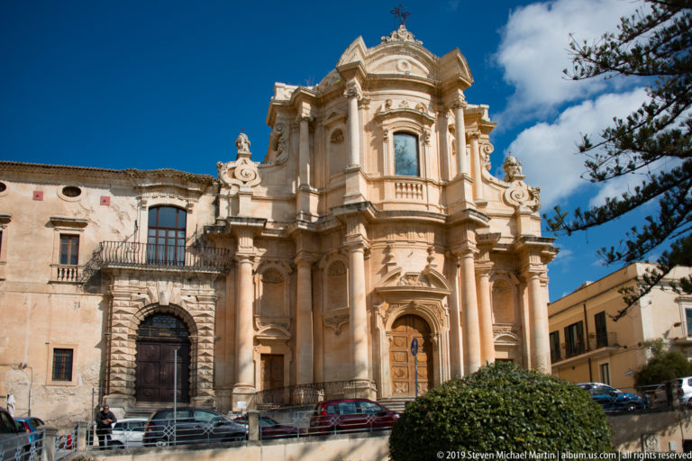 Elaborate curving Baroque church façade with giant columns and sculpted ornament beside an older masonry building.