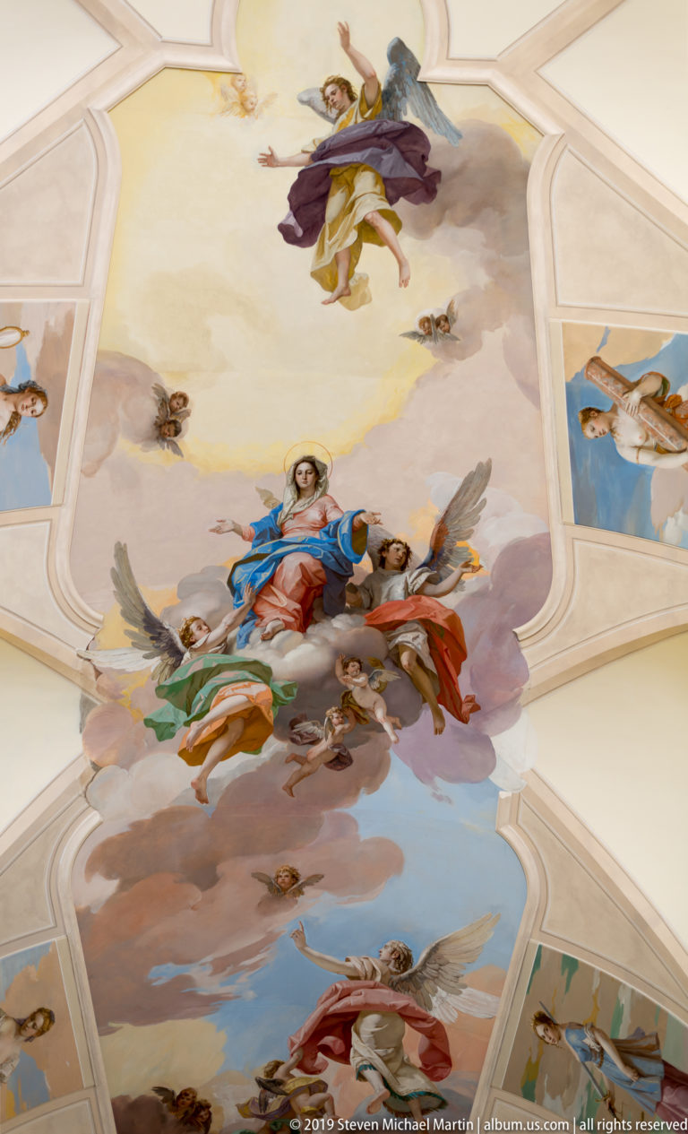Painted church vault with the Virgin in blue and red amid clouds and angels, surrounded by additional ceiling scenes.