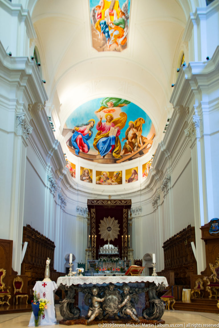 Wide church interior with altar, dark carved front, choir stalls, red IHS hanging, and colorful apse painting above.