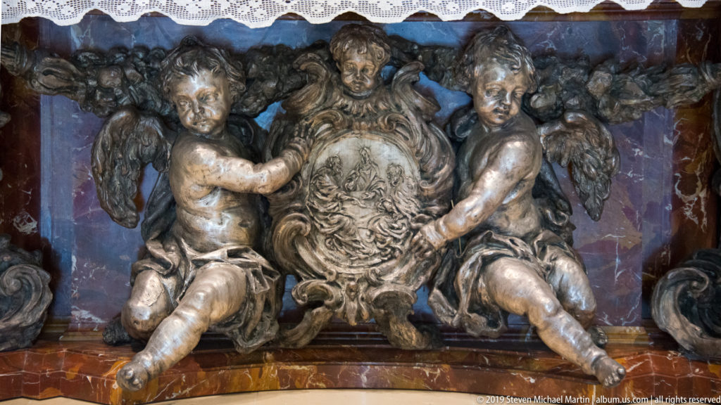 Two sculpted putti support an ornate central cartouche against dark veined marble and dense relief decoration.