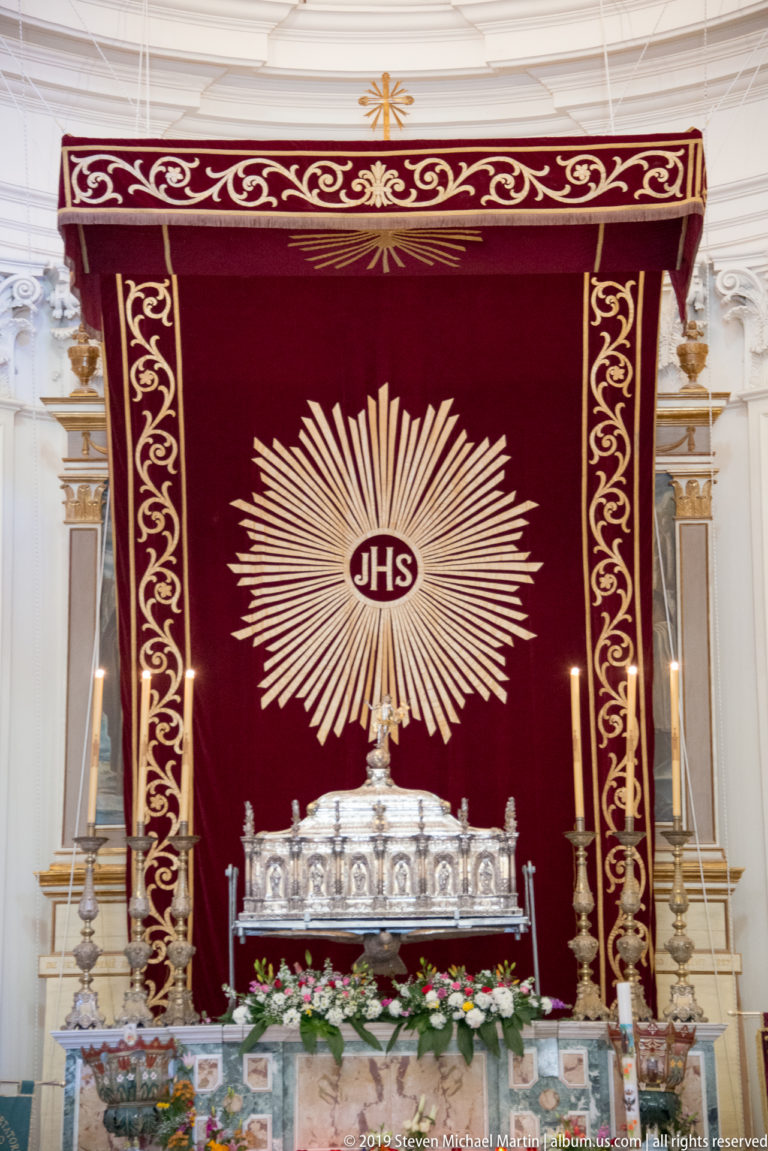 Red altar hanging with radiant gold IHS monogram above silver liturgical objects, candles, and flowers.