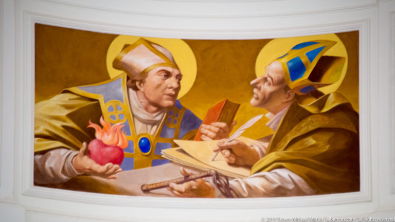 Wall painting of two haloed bishops in blue-and-gold vestments, one holding a red heart-like object and the other writing in a book.