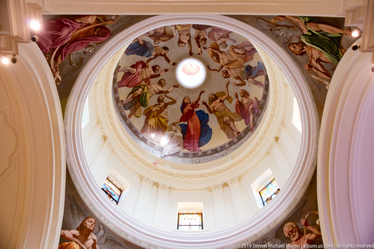 Oblique upward view of a frescoed church dome above a white drum with small windows and broad surrounding arches.