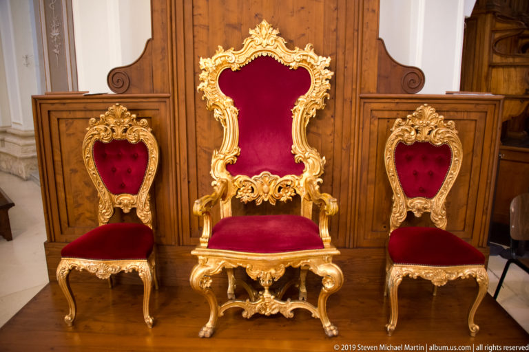 Tall gilded red velvet ceremonial chair centered between two smaller matching chairs against dark wood paneling.