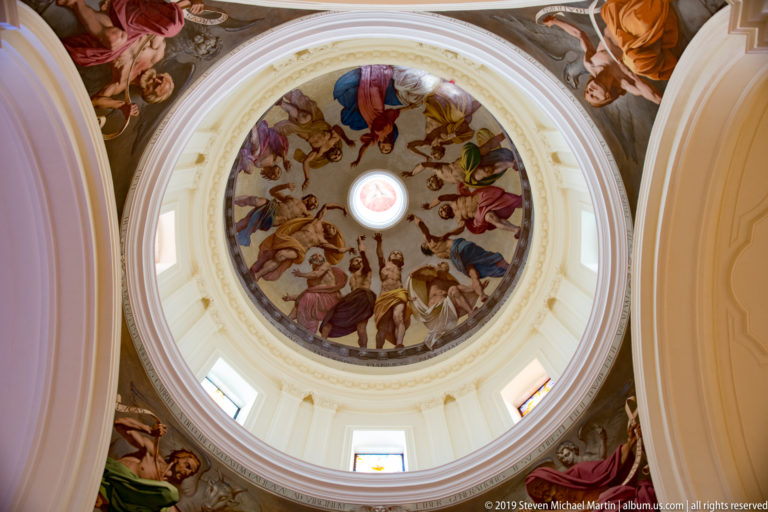 Wide upward view of a white dome with circular fresco, central oculus, drum windows, and painted figures on surrounding pendentives.