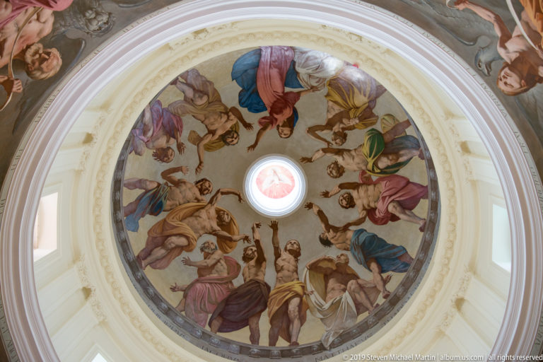 Upward view into a white church dome with a circular fresco of many sacred figures surrounding a bright central oculus.