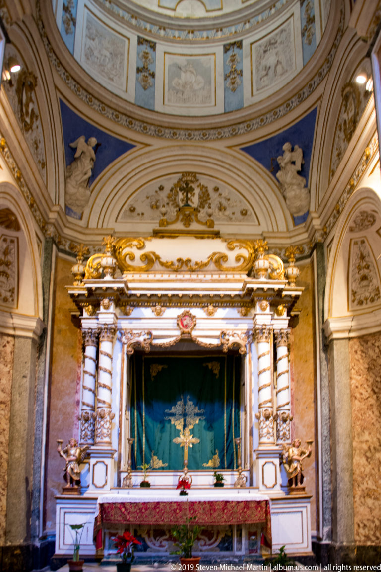 Ornate side altar with paired columns, gilded scrollwork, veined marble, and a crucifix against a dark blue-green backdrop.