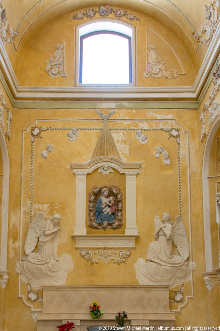 Golden church wall with white stucco scrollwork, two reclining relief figures, and a small central Marian image beneath a window.